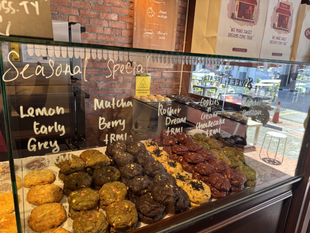 A glass display case at Cookiecrumbs Bangsar filled with assorted thick cookies. Handwritten signs on the glass show flavors like Seasonal Special, Lemon Earl Grey, and Matcha Berry.