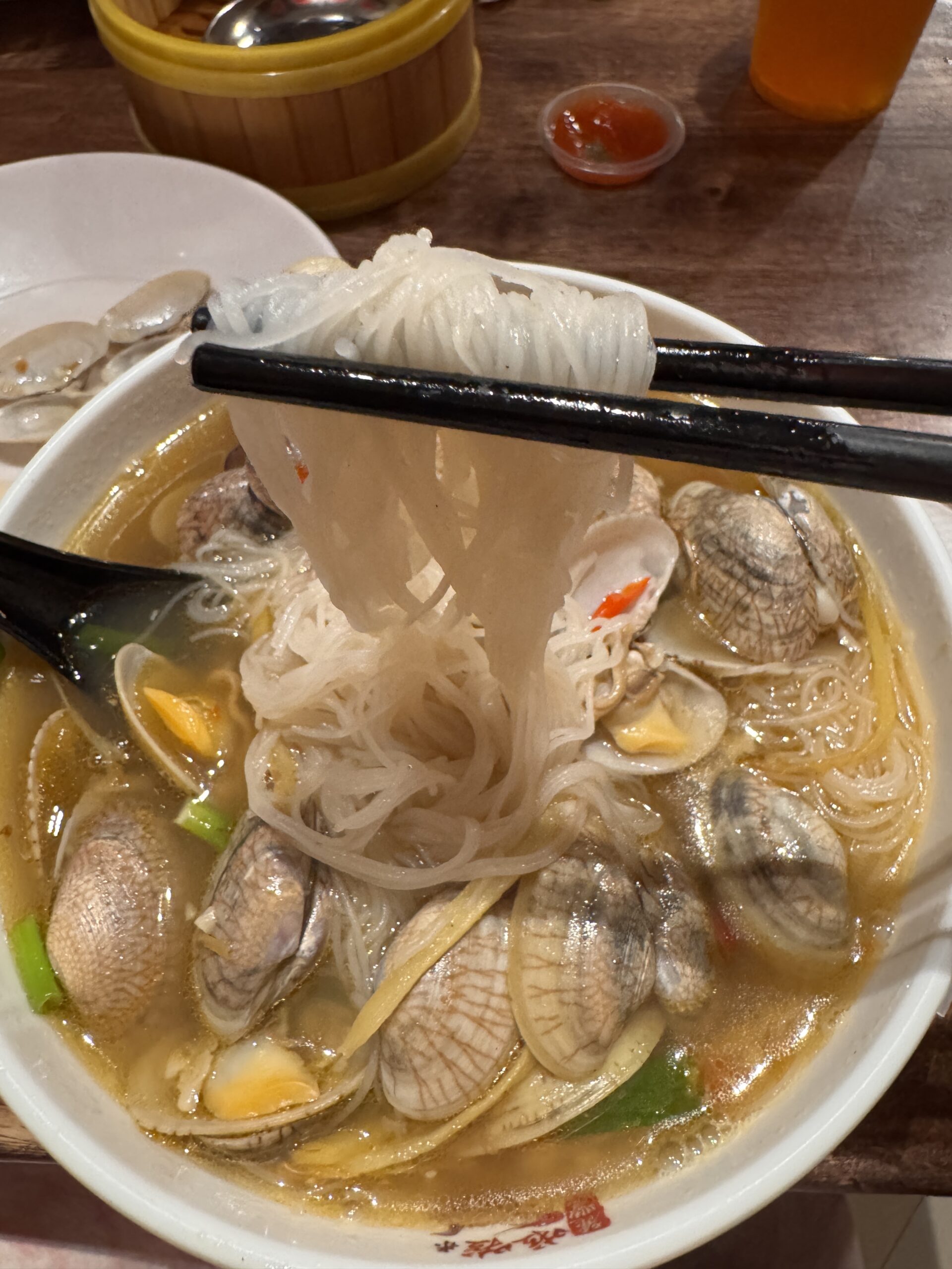 Close-up of thin bee hoon rice noodles lifted from a bowl of lala noodles with clams in clear broth.