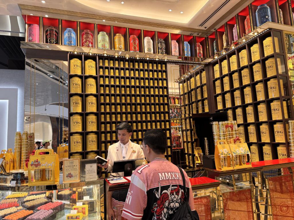 Interior of a TWG Tea store at TRX Mall in Kuala Lumpur, featuring shelves filled with yellow tea tins and a counter where customers are being served.