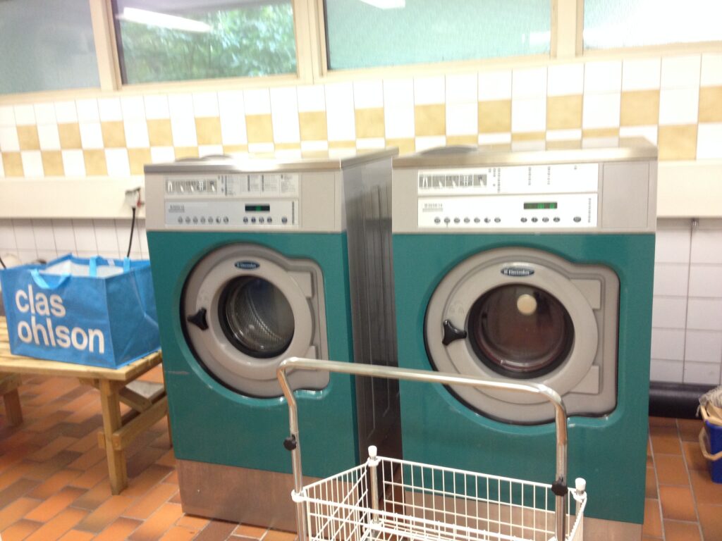 shared laundry room in Sweden with industrial washing machines and dryers, and a blue Clas Ohlson laundry bag placed nearby.