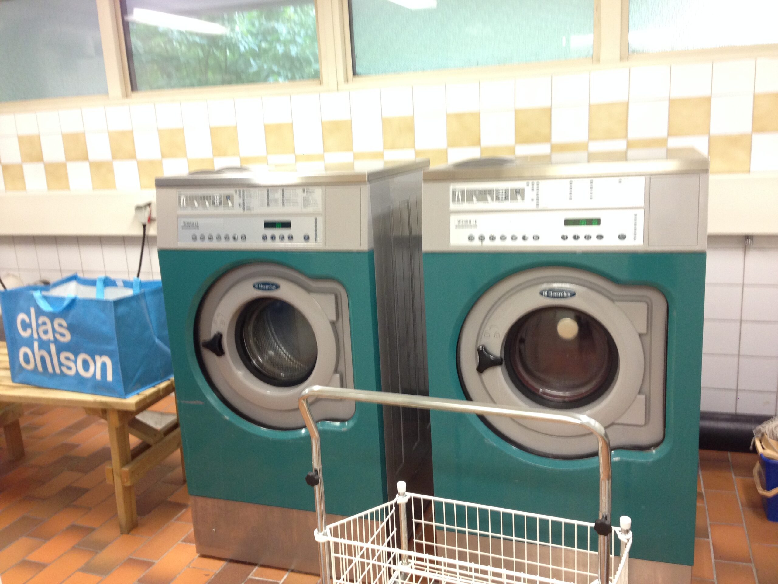 Communal laundry room in Sweden with industrial washing machines and dryers, and a blue Clas Ohlson laundry bag placed nearby.