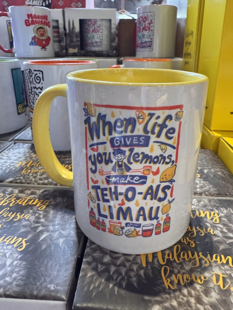 Humorous souvenir mug sold at BookXcess in REXKL, featuring a Malaysia-themed phrase and colourful illustration.