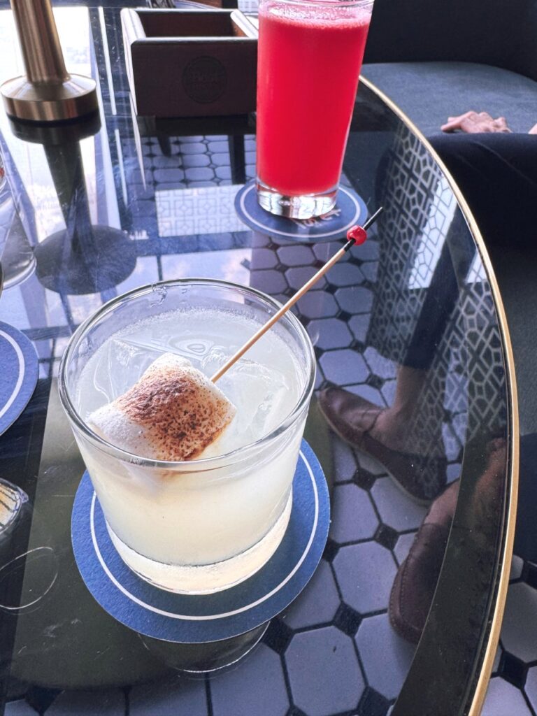 A yellow gin-based "Lee" cocktail with a toasted marshmallow next to a tall glass of fresh red watermelon juice on a bar table at JANN.