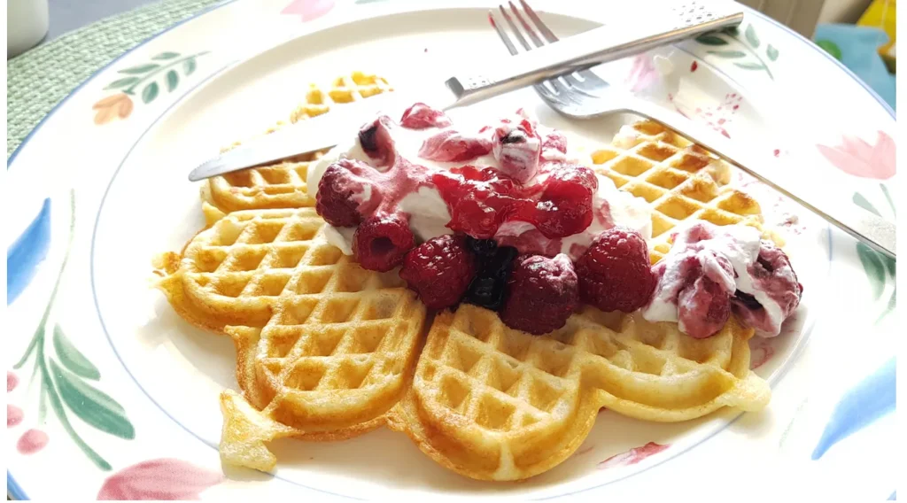 A crispy heart-shaped Swedish waffle topped with whipped cream and fresh raspberries on a floral plate.